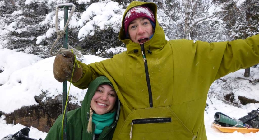 Two people wearing winter gear stand in a snowy landscape and smile for the photo